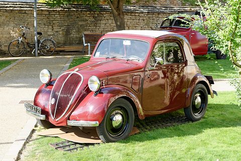 Red small saloon on a lawn before a stone wall, bicycles and old cars behind.