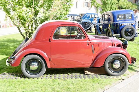 Red small saloon side-on on grass, black wheels, a blue car behind. Opens a larger view