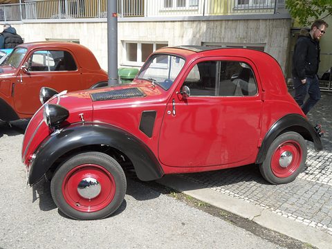 Red small saloon with black wings and red wheels on cobbles, a man walking behind. Opens a larger view