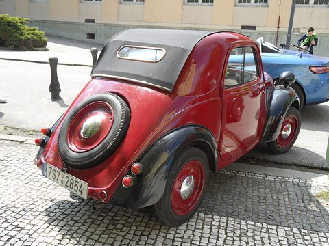 Red small car from the left rear with a black fabric roof and a spare wheel on the tail. Opens a larger view