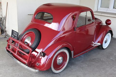 Red small saloon from the left rear with a chrome grid around its spare wheel and whitewall tyres. Opens a larger view