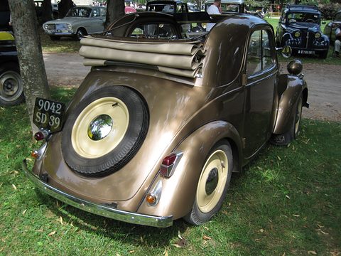 Bronze-brown small car from the left rear, its fabric roof rolled up and a spare on the tail, on grass. Opens a larger view