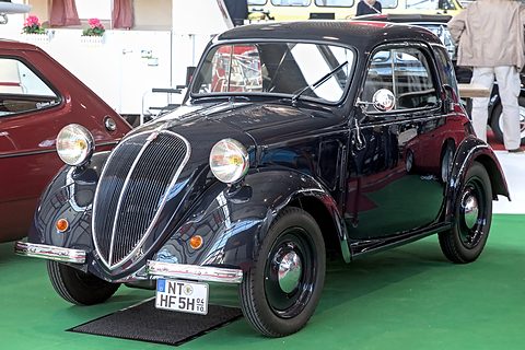 Black small saloon with a tall chrome-edged grille on green show carpet. Opens a larger view