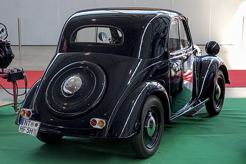 Black small saloon from the left rear, a spare wheel on its rounded tail, on green carpet. Opens a larger view