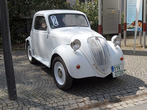 White small saloon with a number 2 on the windscreen and a tall grille, on cobbles under trees. Opens a larger view