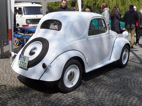 White small saloon from the left rear with a spare wheel on the tail, visitors behind. Opens a larger view