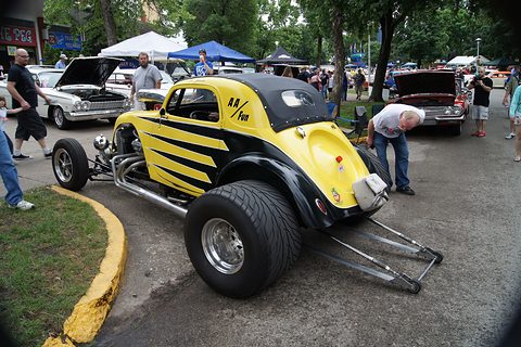 Yellow and black hot rod with a Topolino body, fat rear tyres and a wheelie bar, among show visitors. Opens a larger view