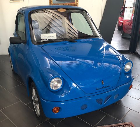Blue retro-styled microcar with round headlamps and a domed bonnet on dark tiles in a museum hall. Opens a larger view