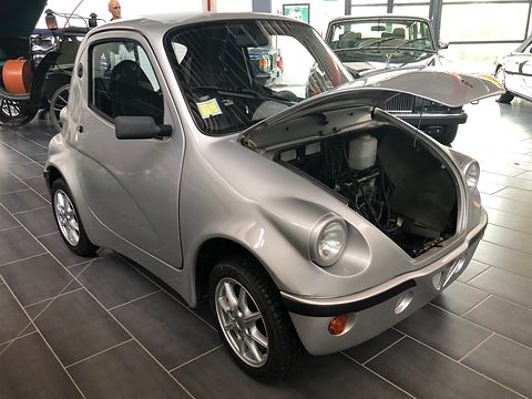 Silver retro-styled microcar with its front lid raised over the motor compartment on a tiled floor. Opens a larger view
