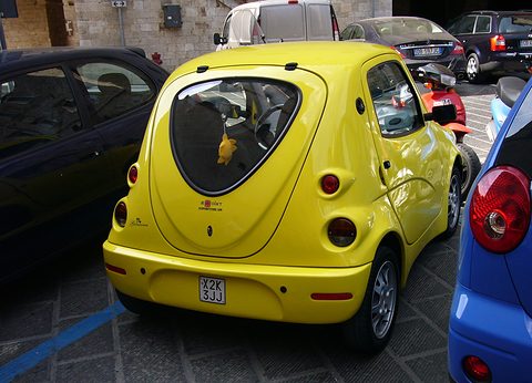 Rear three-quarter of a rounded yellow microcar with a spare wheel cover on the tail between parked cars. Opens a larger view