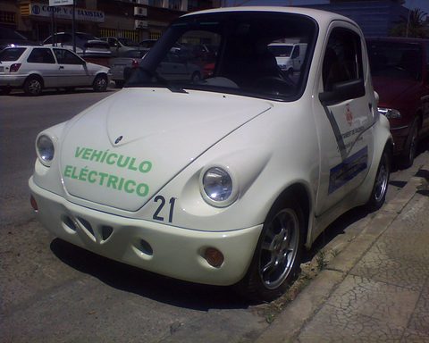 A small rounded white two-door car with green VEHÍCULO ELÉCTRICO lettering and a number 21 on its bonnet, parked at a kerb. Opens a larger view