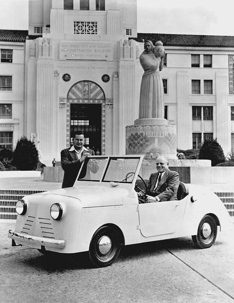 A small white open car with a slatted nose, one man at the wheel and another standing by its windscreen, a large civic building behind.