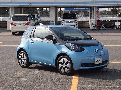 Light-blue city car with a black bonnet and an eQ plate in a car park, minivans and pit buildings behind. Opens a larger view