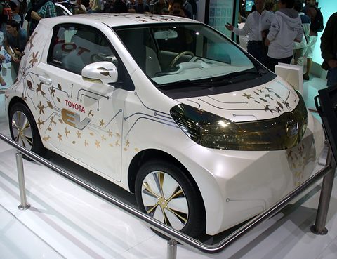 White city car decorated with gold flowers and circuit lines, behind a rail on a show stand, visitors behind. Opens a larger view