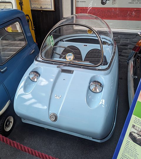 Pale blue bubble car facing the camera, its clear dome closed, beside a blue van. Opens a larger view