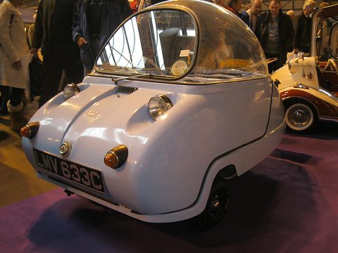 Pale blue bubble car with twin round headlamps and its dome closed, visitors behind. Opens a larger view