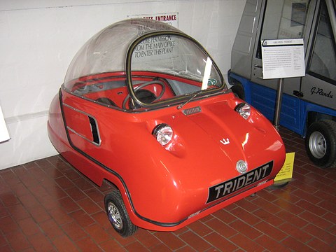 Red bubble car head-on with its dome closed, an older van and a sign behind. Opens a larger view