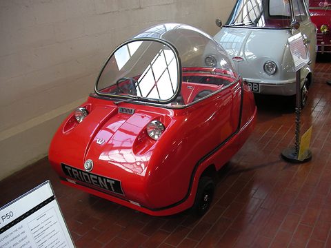 Red bubble car with its dome closed on a tiled floor, a pale microcar behind.