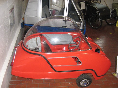 Red bubble car from behind with its curved side moulding, small cars parked beyond. Opens a larger view
