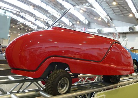 Red bubble car side-on on a metal ramp under an exhibition-hall roof.