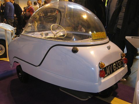 Pale blue bubble car from behind with its dome closed, show visitors standing around it. Opens a larger view