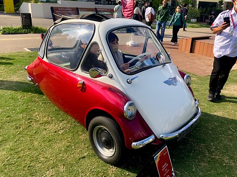 Red and white bubble car on grass with a driver inside, a man standing beside. Opens a larger view