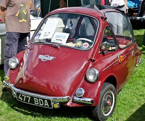 Dark red bubble car on grass with a display card on its dashboard, a man beside. Opens a larger view