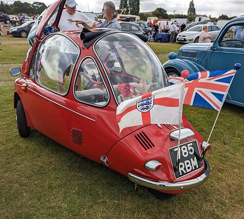 Red bubble car with its front door swung up and English flags fixed to the bumper, on grass. Opens a larger view