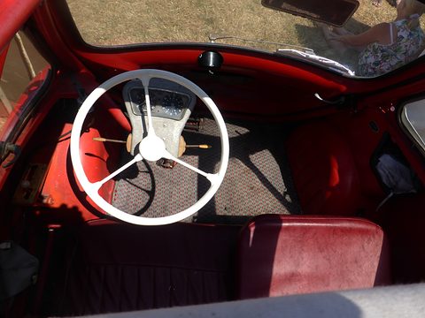 White three-spoke steering wheel over a red interior and patterned carpet, seen from above. Opens a larger view