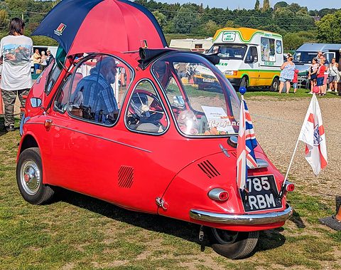 Red bubble car in profile on grass with a red umbrella and flags, a driver seated inside. Opens a larger view