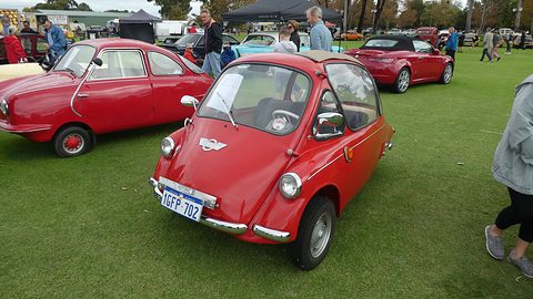 Red bubble car on a playing field beside another red microcar, spectators behind. Opens a larger view