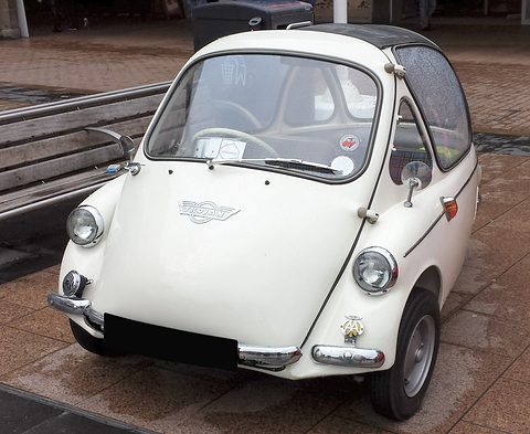White bubble car seen from the front on red paving beside a bench. Opens a larger view