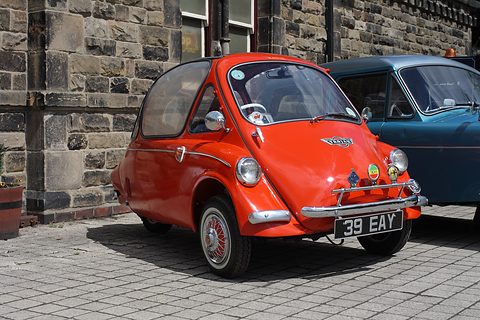 Orange-red bubble car with a badge bar on paving in front of a stone wall. Opens a larger view