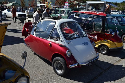Red and white bubble car on tarmac with its roof rolled back, other cars and visitors behind. Opens a larger view