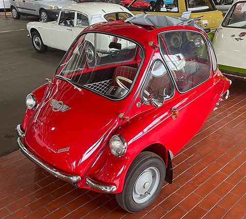 Red bubble car with a tartan seat on a brick museum floor, other small cars behind. Opens a larger view