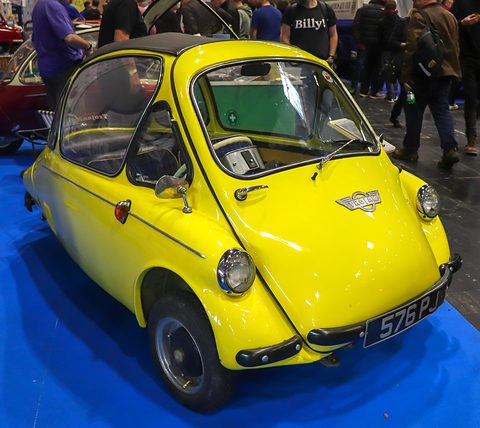 Yellow bubble car on a blue show carpet with visitors crowding behind. Opens a larger view