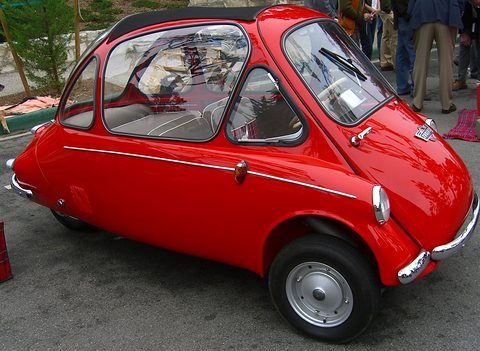 Red bubble car in side view on tarmac with people behind. Opens a larger view