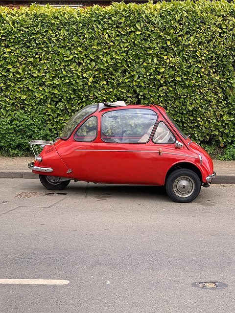 Red bubble car in side view at a kerb in front of a tall green hedge. Opens a larger view