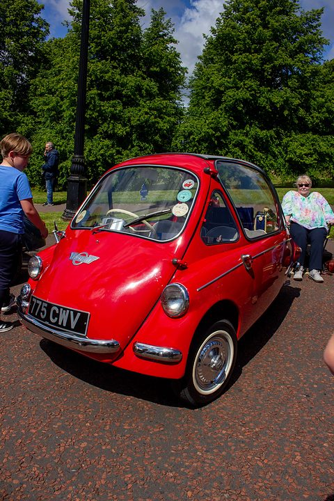 Red bubble car on a red gravel path under trees, spectators at the sides. Opens a larger view