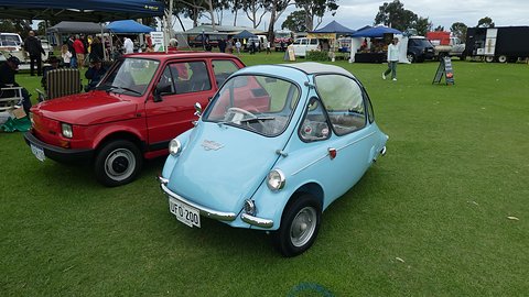 Pale blue bubble car on grass beside a red Fiat, tents and people behind. Opens a larger view