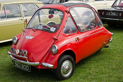Red bubble car with chrome club badges on its bumper, on grass by a cream car. Opens a larger view