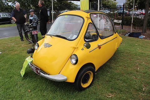 Yellow bubble car parked on grass, onlookers and a bicycle behind. Opens a larger view