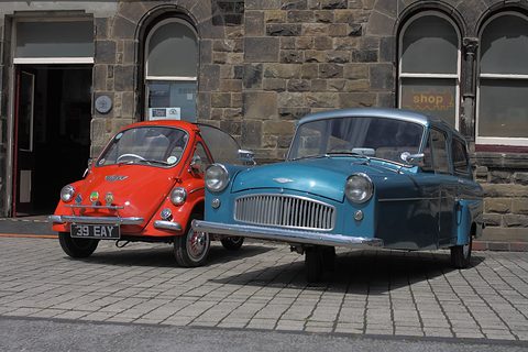 Blue three-wheel saloon and a small red bubble car parked on setts in front of a stone building. Opens a larger view