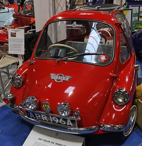 Red bubble car with extra lamps and club badges, on a blue show stand. Opens a larger view