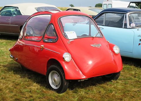 Red bubble car on long grass with a card on its windscreen, a pale blue car behind. Opens a larger view