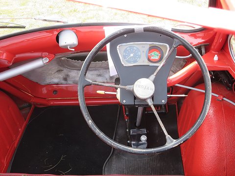 Looking down on a three-spoke steering wheel with a small instrument panel and red interior. Opens a larger view