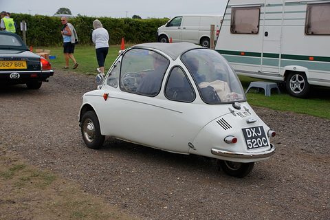 White bubble car seen from the rear right on gravel with a caravan behind. Opens a larger view