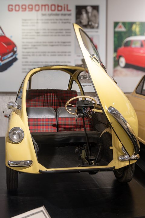 Yellow bubble car with its door raised, tartan seat inside, museum boards behind. Opens a larger view