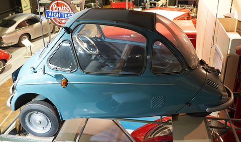 Blue-green bubble car with a black roof on a raised stand, petrol signs behind. Opens a larger view