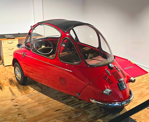 Red bubble car on a wooden floor in a white-walled museum room, seen from the rear right. Opens a larger view